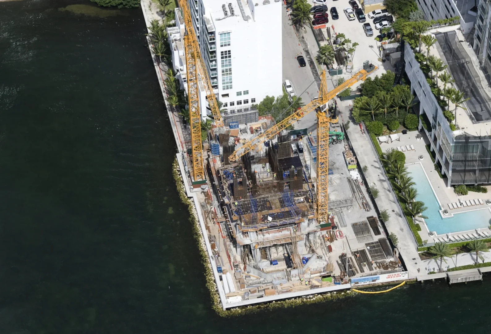 Yellow cranes stand above the Villa Miami construction site beside a body of water and urban buildings