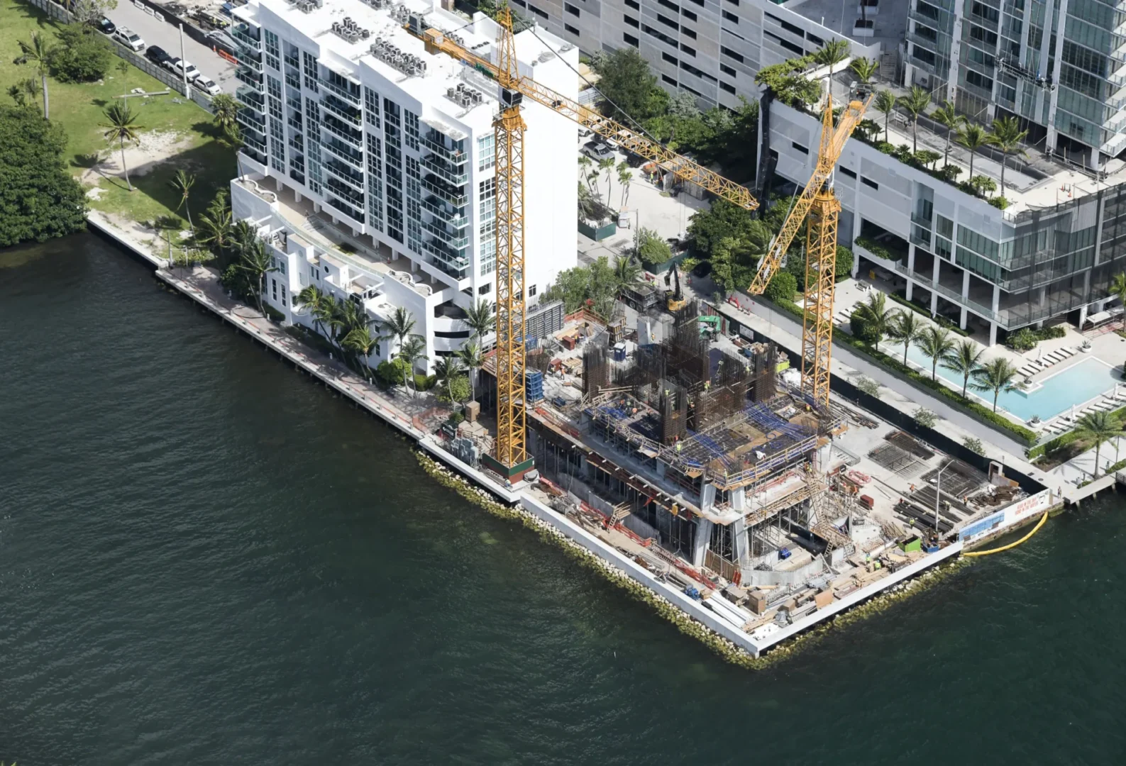Two yellow construction cranes rise above the Villa Miami building site next to waterfront structures and a swimming pool
