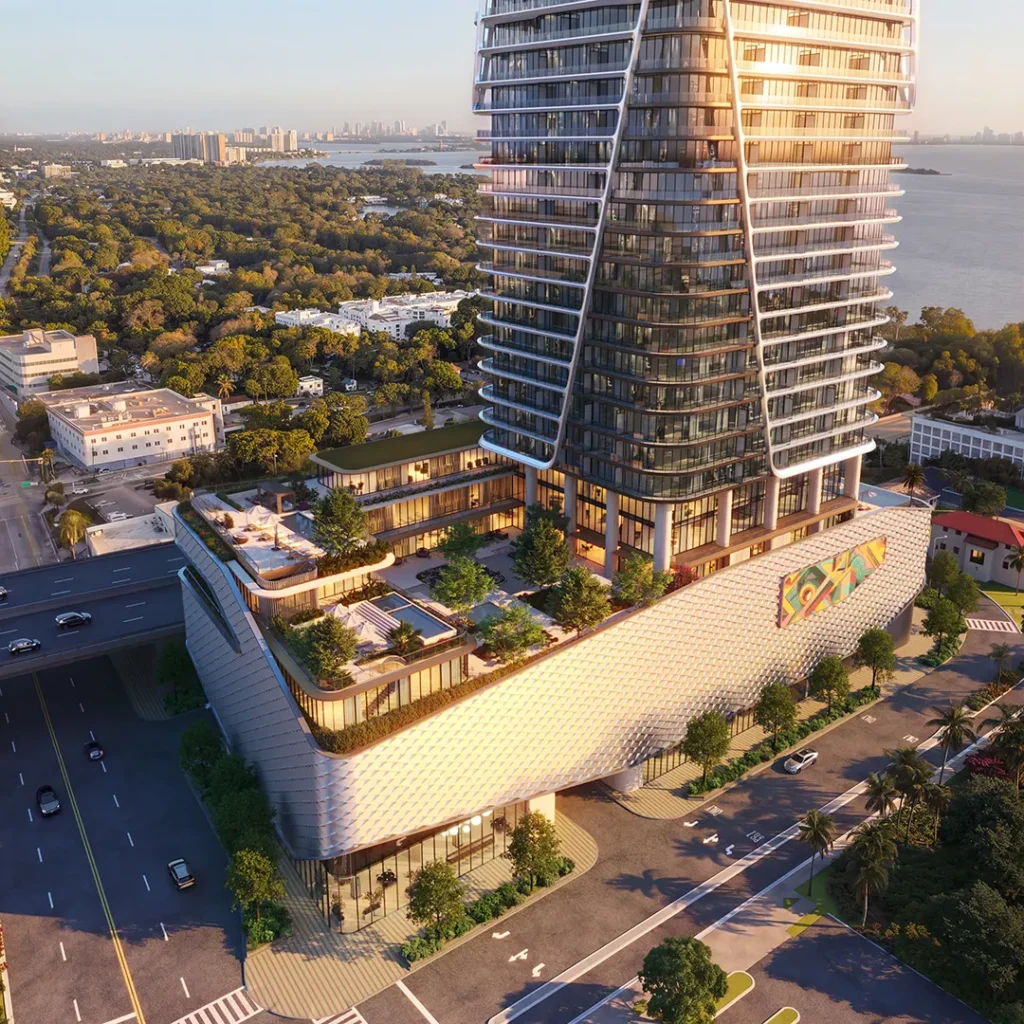A tall glass skyscraper with tiered terraces stands beside an urban street, trees, and a body of water