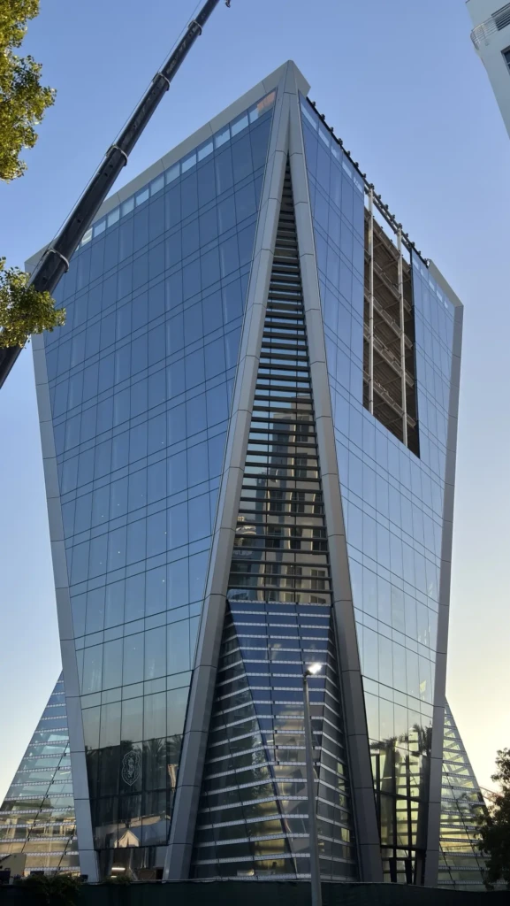 An angular glass building from the Safra 3050 Aventura project stands against a blue sky near a crane arm and green leaves