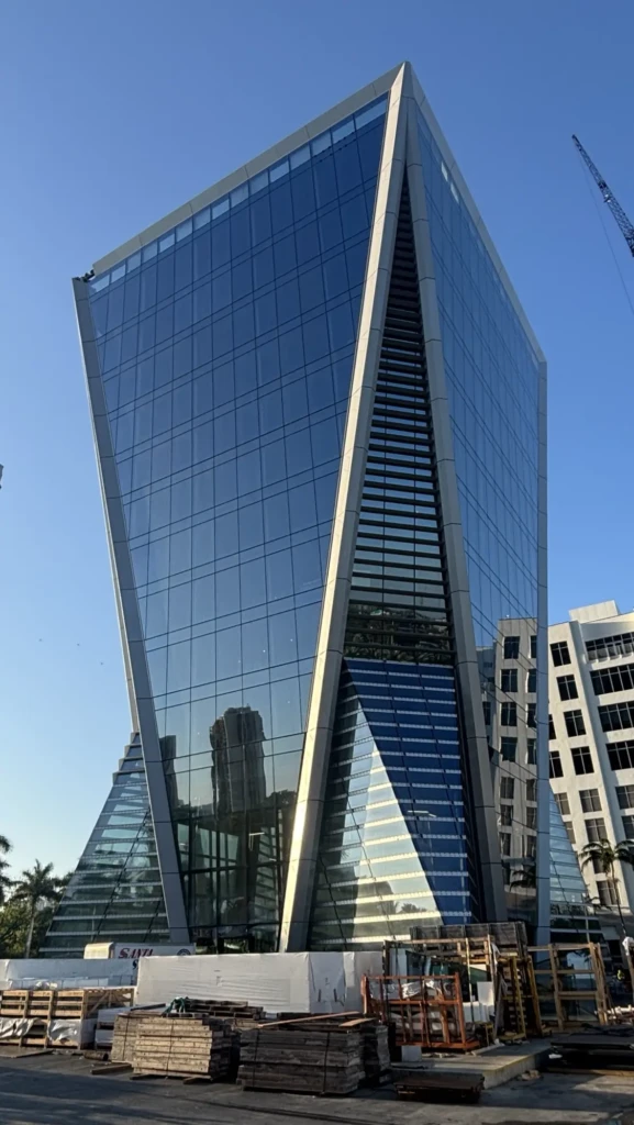 An angular glass building for Safra 3050 Aventura stands behind wooden pallets and construction materials under a blue sky