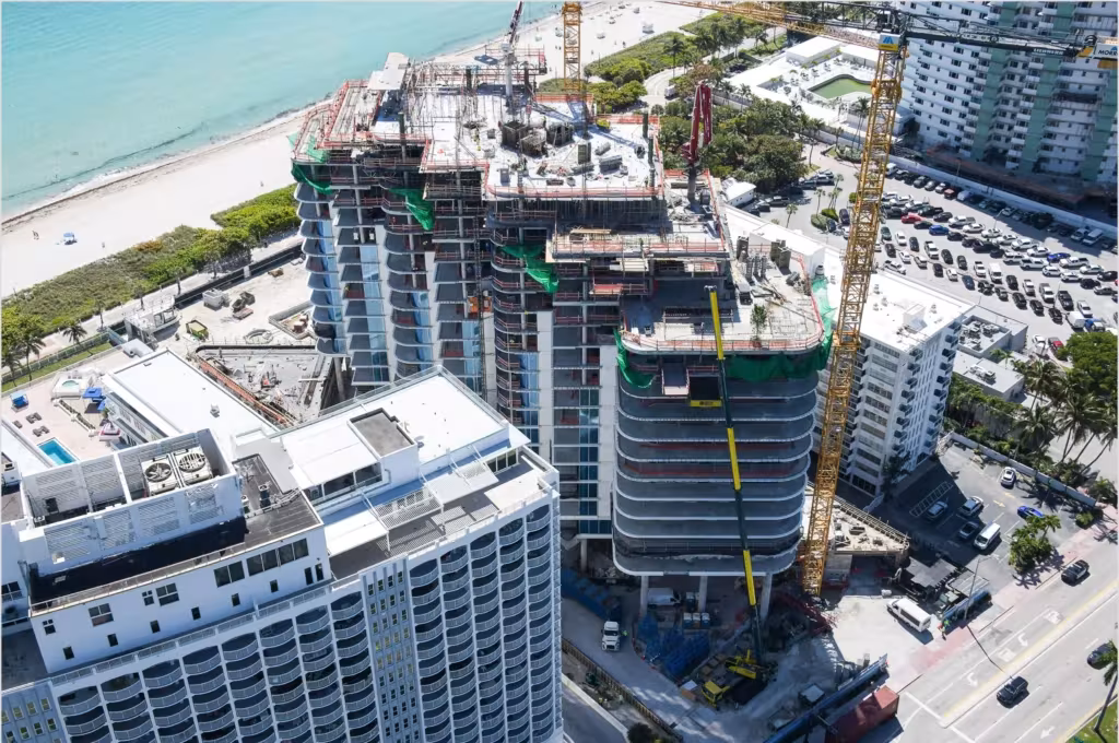A partially constructed high-rise at The Perigon features green safety netting and a tall yellow crane near a beach