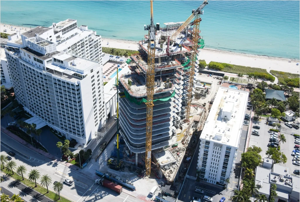 Construction cranes tower over the building under construction at The Perigon near white high-rises and an ocean beach